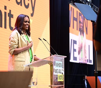Photo of Michelle Obama, wearing green top and tan suit, standing behind a clear podium on stage at the Culture of Democracy Summit.