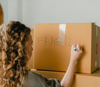 A woman with curly strawberry blonde hair writes “fragile” with a marker on a cardboard moving box.