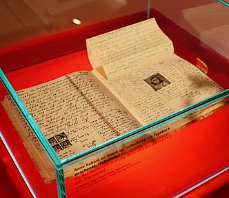 An open book with handwriting and a photo of Anne Frank sits in glass display case on a red platform.