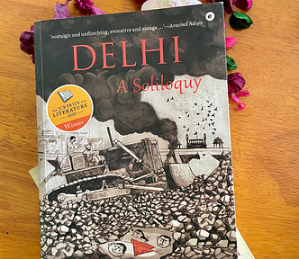 Image of the book, Delhi: A Soliloquy by M Mukundan lying on the table with dried flowers and another book, City of Djinns by William Dalrymple.