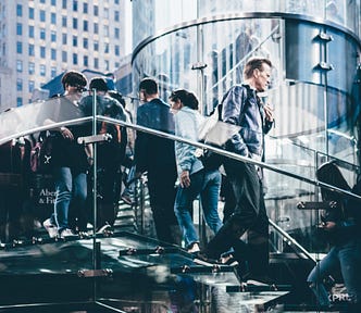 People walking on outside glass staircase in city surrounded by tall buildings