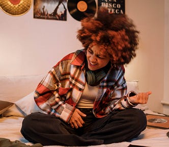 A woman with a multi-colored afro rocking out while sitting on her bed.