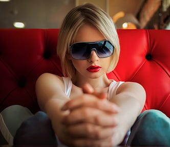 girl with short blonde hair wearing sunglasses and red lipstick with hands clasped in front of her