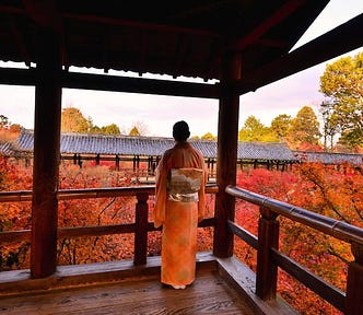 A kimono-clad visitor stands on a wooden veranda at Tofuku-ji Temple in Kyoto during the autumn foliage season, looking out over brilliant red and orange maple leaves. The covered temple walkway frames the scene with dark wooden pillars and railings, while tiled roofs and trees in late-afternoon light stretch across the background. The image captures the quiet beauty of fall in Japan, with the traditional architecture and vivid koyo colors creating one of Kyoto’s most iconic seasonal views.