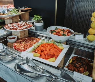 A hotel buffet displaying neatly arranged prepared foods, including sliced salmon, salads, sauces, and plated dishes under controlled conditions, emphasizing abundance, uniformity, and visual order.