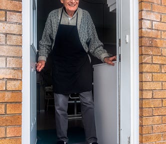 An elderly man in an apron standing at the open external door of his house