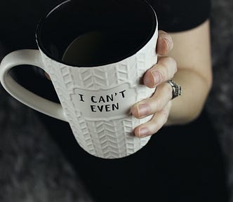 A woman’s hand holding a white mug on which the words “I can’t even” are written in black capitals.
