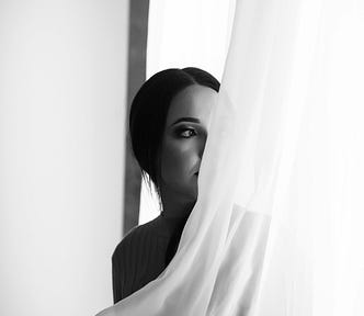 A woman standing by a window, partially veiled by a sheer curtain, holding herself in soft, introspective light.
