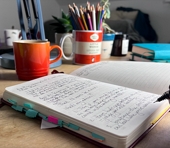 An open notebook, filled with words on the left hand page, with a hand holiding a pen poised to write.