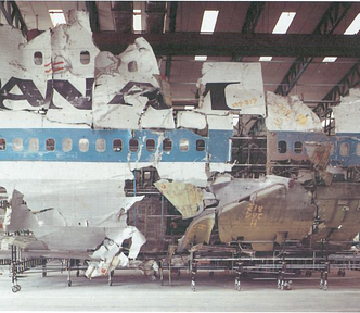Color photograph showing damaged side of airplane with PAN AM logo, housed in a large building.