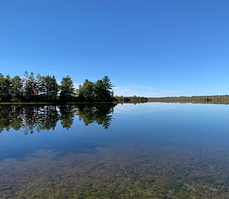 A picture of a calm and clear lake in Maine with the reflection of trees in the water.