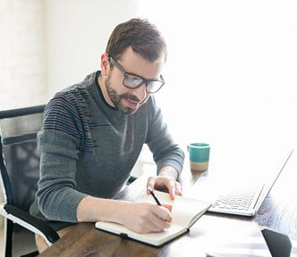 A man writes in his notebook while working at a desk with a laptop.