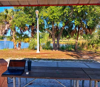 A park table in the shade with a laptop, mouse and drink with water and fountain in the background and a squirrel burrying its nut in Estero park in Florida