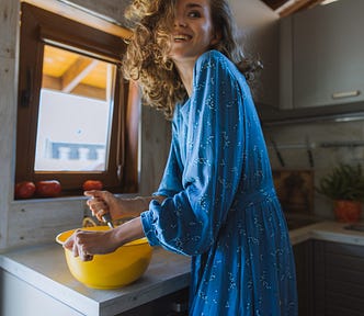 A woman with a beautiful smile and long blue dress. Her face is half covered with her flowing hair. She looks happy and is cooking something.
