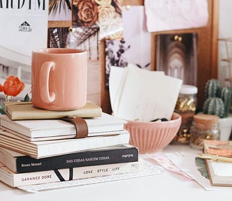 A stack of books and journals sits on a white desk with a pink mug on top of the stack. The desk is covered in a variety of items, including a cactus plant, small pink bowl, and papers. There is a corkboard in the background mostly obscured by photos of flowers and architecture pinned to it.