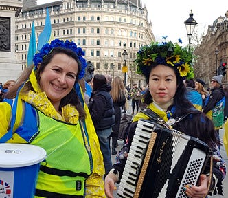 2 ladies wearing flower headdresses, one holding blue charity collection, the other an accordion
