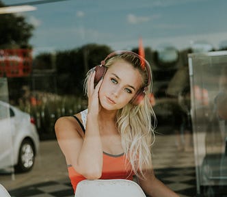 Young woman, model, beautiful face, long blonde hair, seen through a window, seated,listening to music, wears orange top & denim shorts, window reflects the street behind the photographer