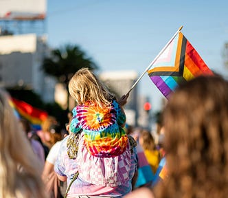 A young child wearing a tie-dye shirt and holding the Pride flag sits atop their parent’s shoulders at a Pride parade