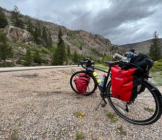 The author’s bicycle, on the Cameron Pass over the Rocky Mountains. It has two red pannier bags over the rear wheels and two red pannier bags over the front wheels; in the background are mountains and a cloudy sky