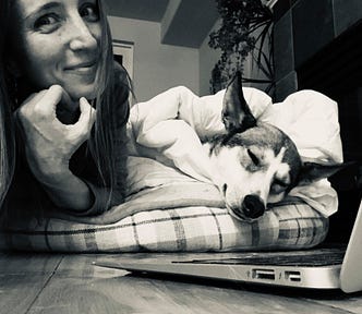 A black and white photo of the author smiling and her sleeping dog in front of her laptop