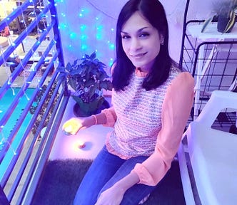 A West Indian Girl with mid-length black hair is holding a diya on Diwali night. She’s wearing jeans and an orange blouse. She also has a smile on her face. In the background, there is a potted plant and some white chairs.