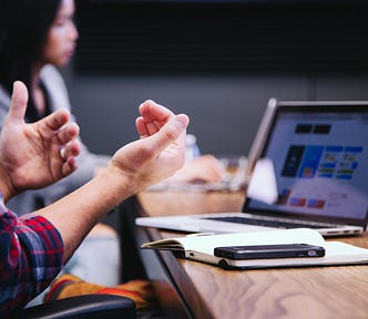 A man gesturing with his hands in a virtual meeting, joined at a conference table by coworkers in the background also on their laptops