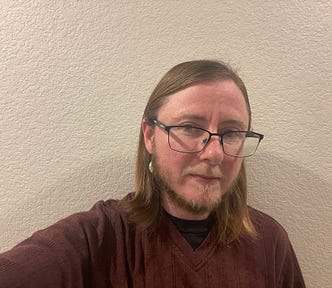 The author is wearing a maroon shirt and is standing in front of a white textured wall. He is wearing black-rimmed glasses, a dangly earring, and has a beard that is gray and brown.