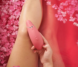 Photo shows a woman’s legs in a bathtub with pink water and pink flowers. In her hand, she holds a pink adult toy with a tapered end, the toy resting on her knee.