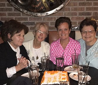 Snapshot of 4 women sitting at a table in a restaurant, in front of a partially eaten birthday cake.