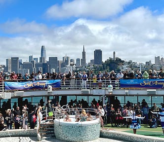 Image of a crowded group of guests and crew dancing and celebrating on the pool deck (level 15) and upper deck (level 16) of the Ruby Princess cruise ship with the San Francisco skyline in the background below a bright blue sky and fluffy white clouds. Two large banners hanging from the guardrails read “Sail Away” party.