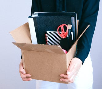 Torso of a female in white slacks and a navy shirt holding a cardboard box full of office supplies