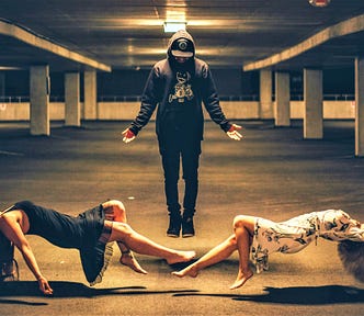 man in dark clothes and hat between two women on their backs levitating in the air in parking garage