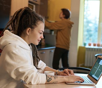 Smiling woman looking at her laptop. She sitting at a table in the kitchen while a man reaches for the microwave in the background.