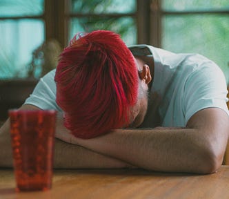 A man with red hair is sitting at a table with his head down, looking depressed