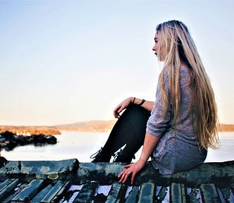 girl with long blonde hair wearing grey top and black pants sitting on wooden dock looking out at water