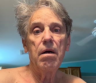 A man (me) with gray hair and a tired expression on his face, sitting in a room with light blue walls and a ceiling fan visible in the background.