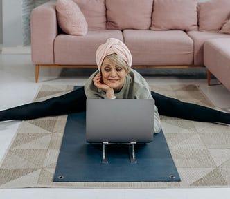 Smiling older woman on a yoga mat with legs stretched out andlooking at a computer screen.