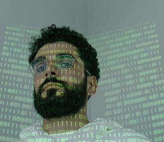 man looks down at camera. his face is covered by a projected screen of binary code (0s and 1s).