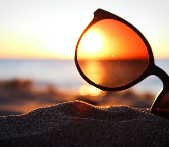 A pair of sunglasses with a black frame sits tilted at an angle on a sandy beach, against a background of sun setting over the ocean.