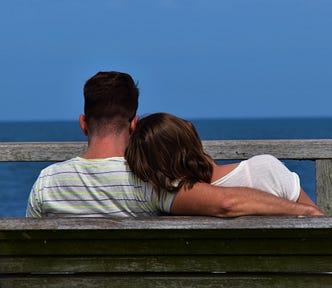 A rear view of a man and a woman sitting on a bench. The man has his arm around the woman’s shoulders, and she has her head resting on his shoulder.