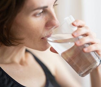 A young woman with short brown hair and wearing a black tank top is drinking a glass of water.