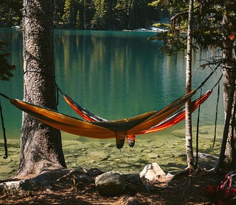 A hammock lays out between two trees. In the background, a crystal clear lake glimmers in the afternoon sun. The image gives a very relaxed and carefree feeling.