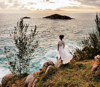 A young woman in a white flowy dress, standing on a cliff, and looking out onto the sea.