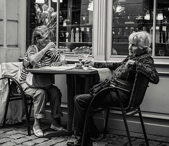 Two women in their seventies sitting at a table outside a cafe. They are not looking at each other.
