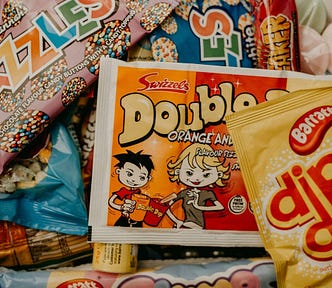 A selection of vintage sweets, including Jazzles, a DibDab, and a sherbert Double Dip. The packaging of the Double Dip depicts a young man offering his candy stick to a young woman, who is reaching for it with a sly grin.