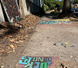 Painted graffiti art on the sidewalk in front of a couple of trees, foreground says, “Get unstuck”, with bright colors of blue, orange, and green. Background graffiti art is a group of unidentified letters. Autumn leaves litter the ground beside the concrete, and another painting in black and red graffiti on a board is propped up against a wall. Art courtesy of my homeless friend Maize, who lived across the street from my apartment, not fully pictured.