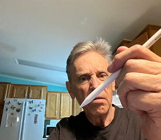An older man in a kitchen holds an Apple Pencil close to the camera with a curious, slightly skeptical expression on his face. Wooden cabinets and a refrigerator with butterfly magnets are visible in the background.