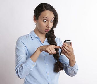 A startled-looking woman with a long, thick braid of dark hair — wearing a long-sleeved blue shirt — touches a smartphone with her right index finger.