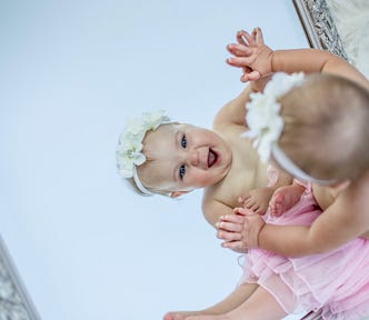 A baby dressed in a pink tutu and a flowery head-band delighting in her reflection in the mirror.