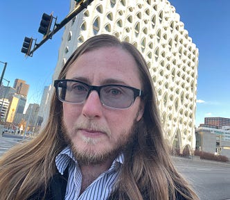 The author has long hair and a beard. He’s wearing sunglasses and a stripped dress shirt with a coat. He’s standing on a street corner in front of a white building with skyscrapers and a streetlight in the background. There are no clouds in the blue sky.